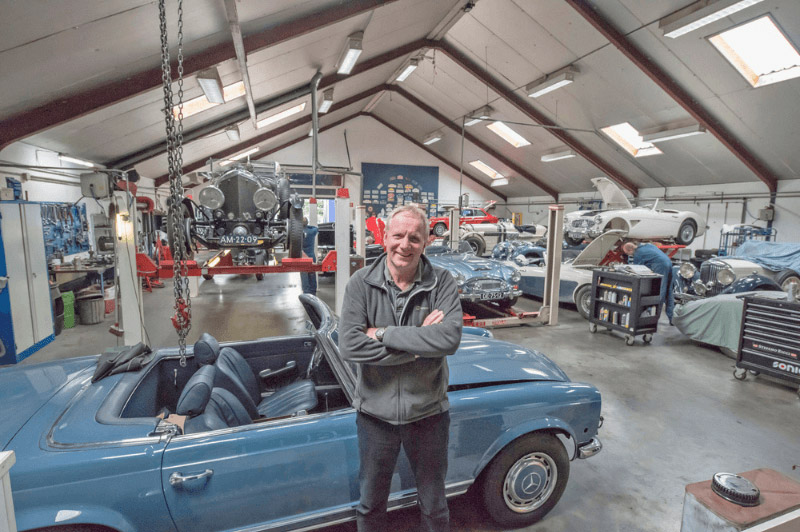 Eine männliche Person mittleren Alters lehnt an einem hellblauen Oldtimer-Cabrio in einer Autowerkstatt.